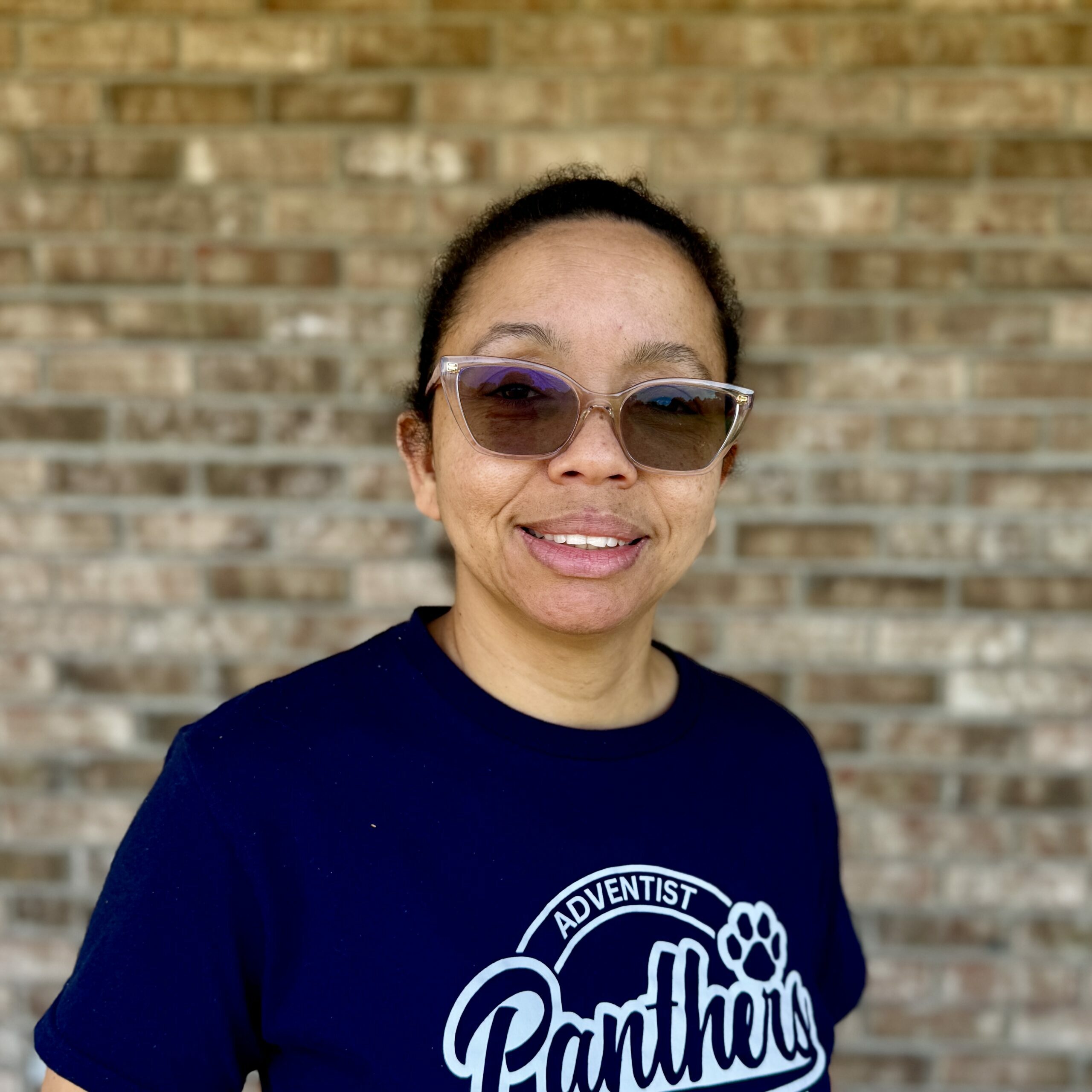 Close-up portrait of a person wearing sunglasses against a brick wall background, smiling slightly.