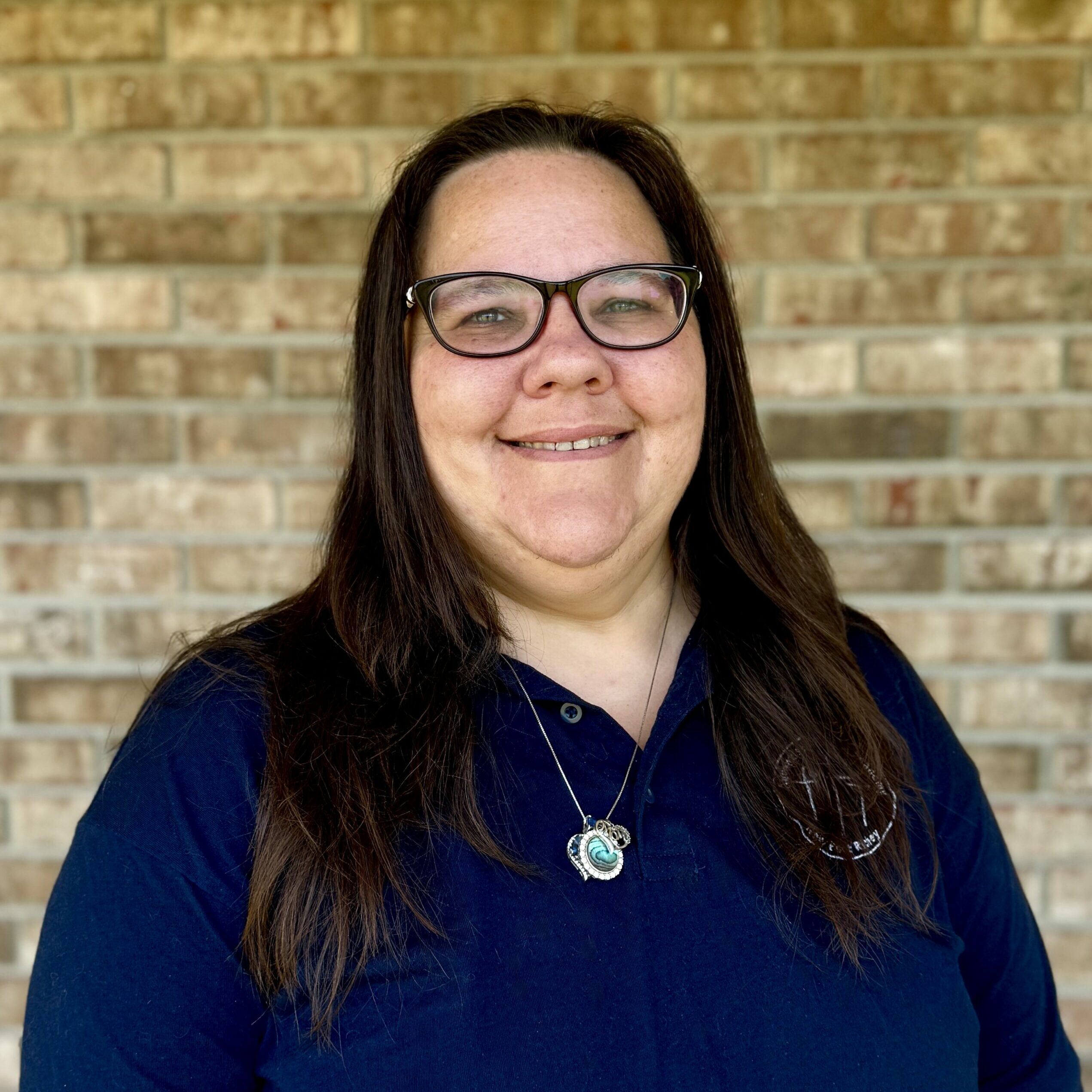 Close-up of a woman with long dark hair wearing glasses and a turquoise pendant necklace, against a brick wall background on a navy top.