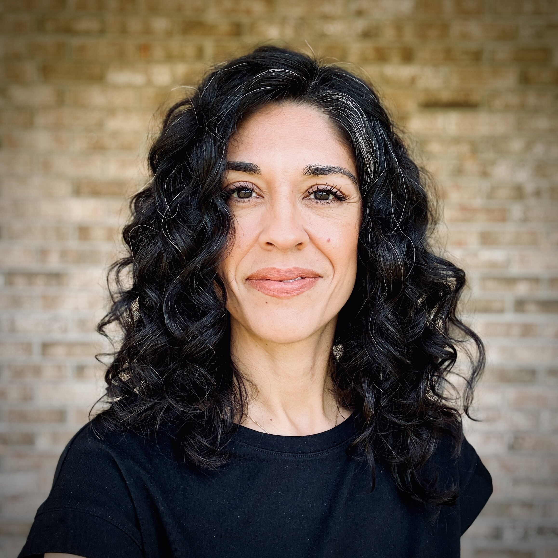 Close-up of a woman with dark curly hair, wearing a black top, with a brick wall in the background.